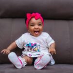 Cute baby with pink headband smiling joyfully on a sofa.