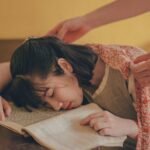 A young woman falls asleep on a book as someone covers her with a blanket.
