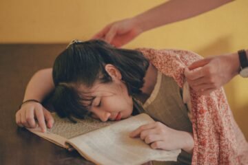 A young woman falls asleep on a book as someone covers her with a blanket.