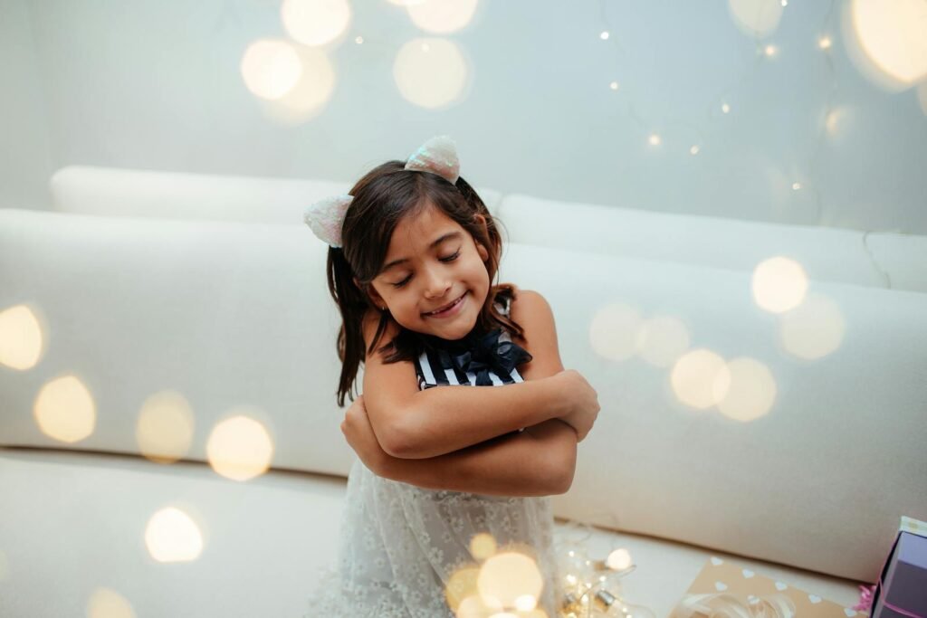 Cute child wearing cat ears, hugging herself amidst glowing fairy lights indoors.