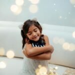 Cute child wearing cat ears, hugging herself amidst glowing fairy lights indoors.